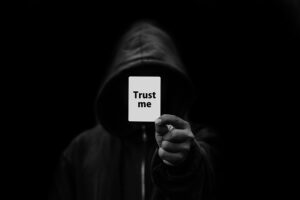 Dark black and white image of a hooded man holding up a card with Trust Me written on it.