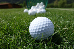 One golf ball on the green, in the foreground in sharp focus, with a pile of other golf balls behind out of focus.